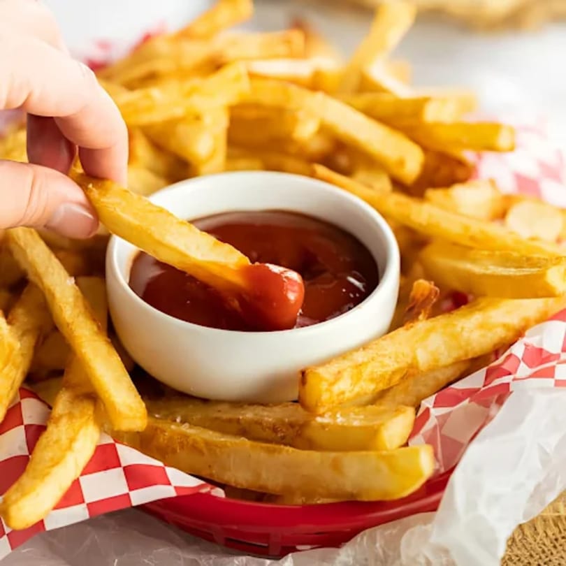 Hand dipping a golden french fry into a white bowl of ketchup, surrounded by more fries.